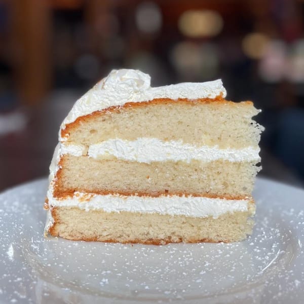A slice of vanilla cake with three layers of white frosting and powdered sugar on a white plate.