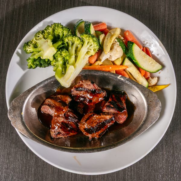 Grilled steak bites in a metal dish with broccoli and mixed vegetables on a white plate.