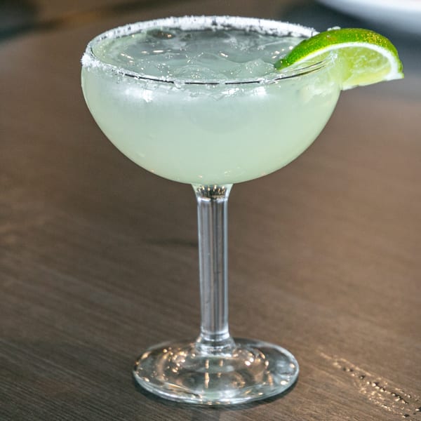 A margarita in a stemmed glass with a salted rim and a lime wedge, on a wooden table.